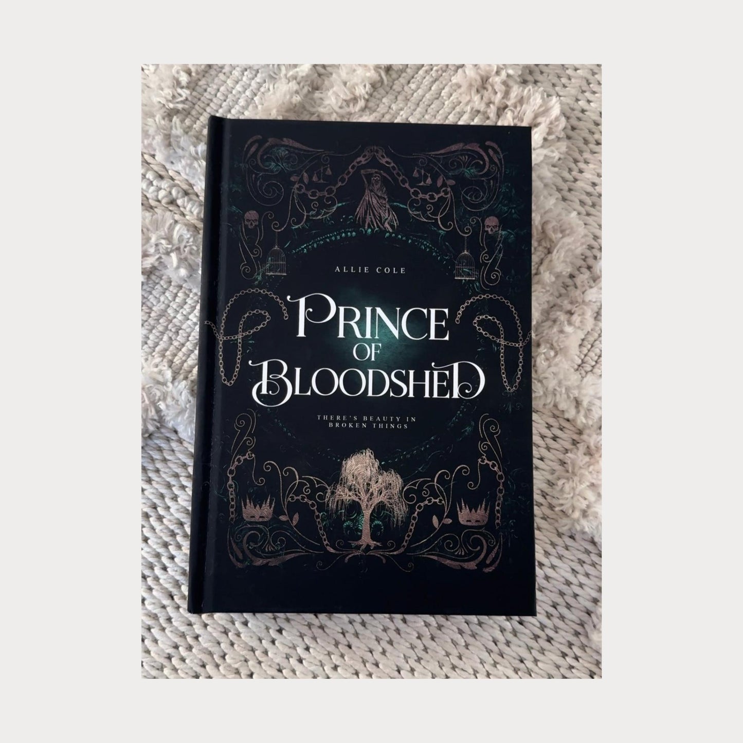 Hardcover edition of Prince of Bloodshed by Allie Cole, a dark fantasy romance novel. The book rests on a textured beige surface, featuring an intricate gothic cover design with chains, skulls, birdcages, and a deep green background. A golden, thorn-covered crown sits at the bottom, and the tagline reads There’s beauty in broken things