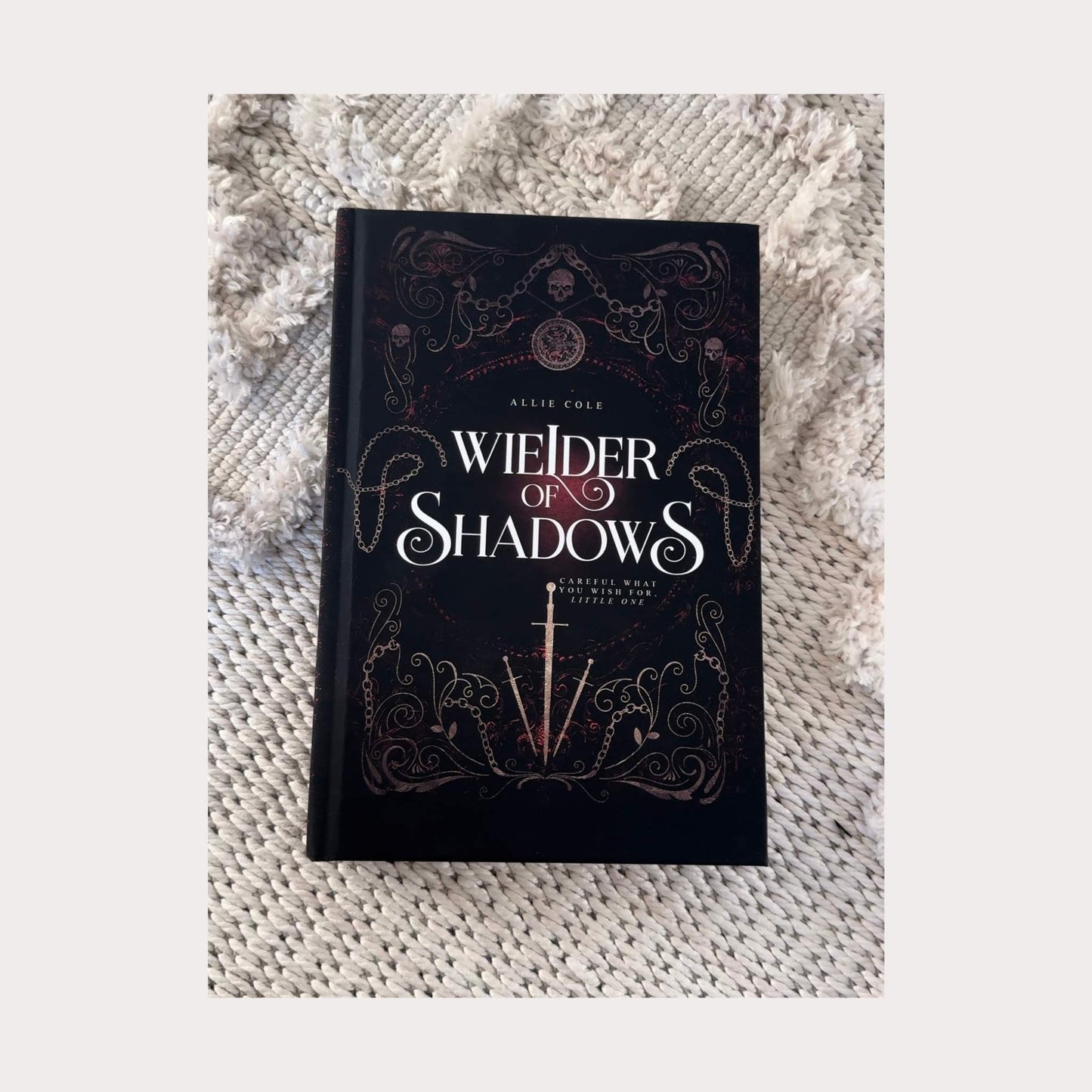 Hardcover edition of Wielder of Shadows by Allie Cole, a dark fantasy romance novel. The book rests on a textured beige surface, showcasing an intricate gothic cover design with filigree, chains, skulls, and a glowing red background. A silver sword is centered beneath the title, with the tagline Careful what you wish for, little one
