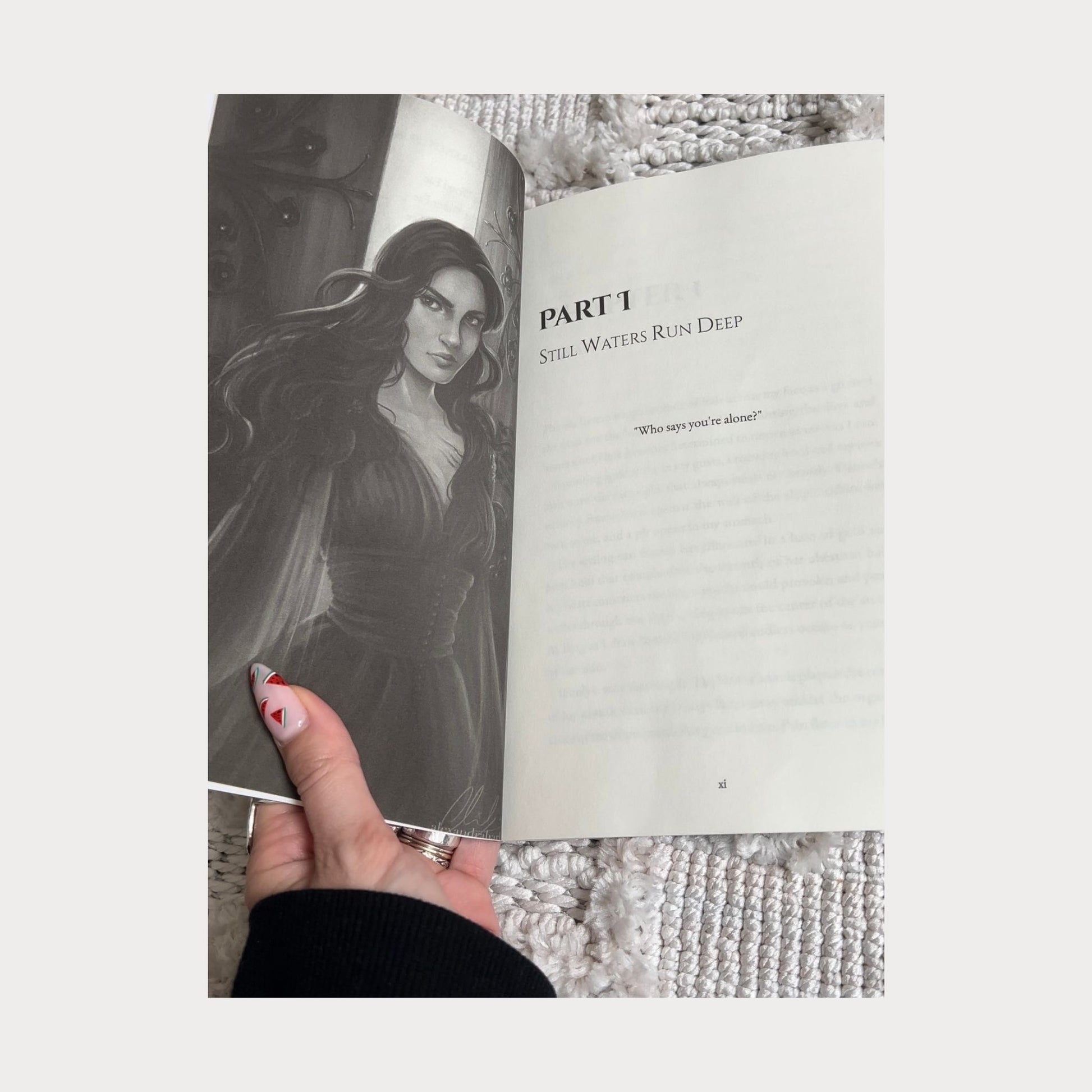 A reader holding an open copy of Daughter of Darkness Prince of Bloodshed by Allie Cole, showing a black and white character illustration on the left page and the start of Part 1 titled Still Waters Run Deep on the right page. The book rests on a textured white surface