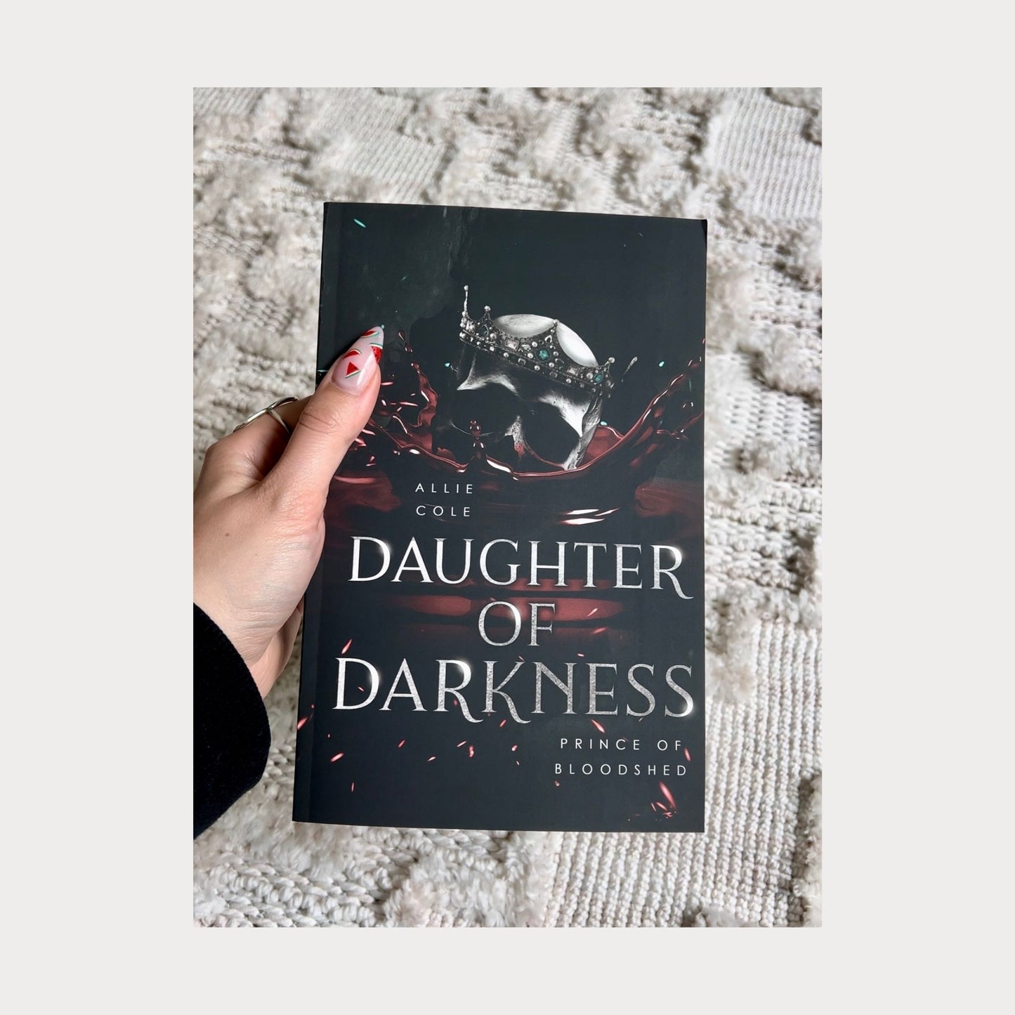 A reader holding a paperback copy of Daughter of Darkness Prince of Bloodshed by Allie Cole, a dark fantasy romance novel. The book cover features a silver skull with a jeweled crown emerging from red liquid against a dark background, held over a textured white surface